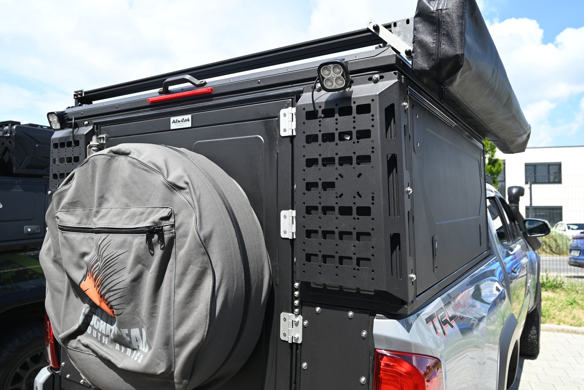 ModCAP Worker zu sehen. Montiert auf einem Offroad Fahrzeug. Mit Geschlossener Hecktür und Seitentüren. Blick auf die Molleplatte neben der Hecktür. Mit montiertem Ersatzrad, Lazerlamps und einer Markise.